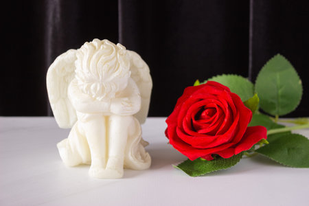 A small sculpture of a baby angel with wings and a red rose on a black background. The concept of funerals, condolences, and mourning.の写真素材