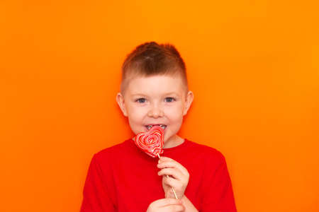 A boy eats a heart-shaped lollipop on an orange background. Fun, sweets for children.の写真素材