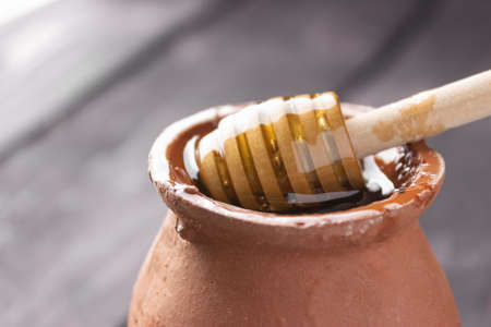Bee honey in a clay pot on a wooden table. Wooden spatula for honey. Side view, close up. Healthy food. Farm, apiary.の写真素材