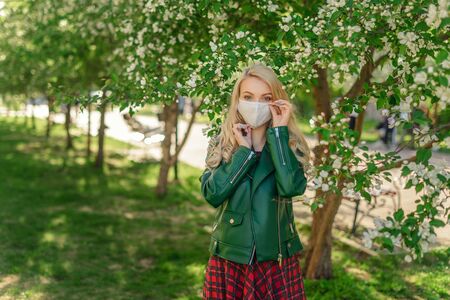 A blonde girl with beautiful eyes stands in the open air near blooming apple trees and adjusts the protective mask against coronavirus on her face.の写真素材
