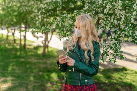 A blond girl in a white medical mask and green jacket holds a white smartphone in her hands and looks at his screen, standing next to a blossoming apple treeの写真素材