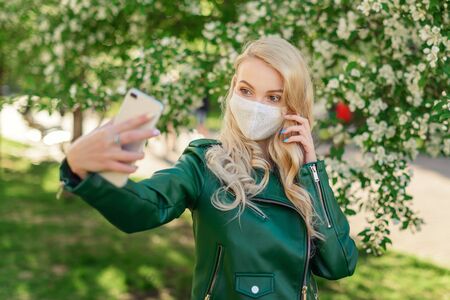 A blonde girl in a white medical mask and a green jacket holds a white smartphone in her hands and takes a selfie, standing next to a blossoming apple treeの写真素材