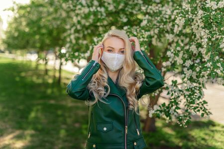 Blond girl in a white medical mask and a green jacket straightens her hair, standing near a blossoming apple treeの写真素材