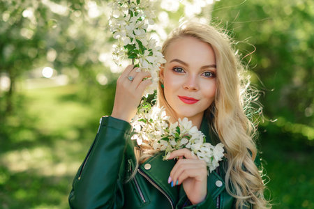 Beautiful blonde girl holding a flowering branch of an apple tree in her hands. Close-up portrait of a young girl with long hair.の写真素材