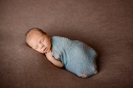 sleeping newborn baby wrapped in gray-blue knitted fabric, lying on its side against a brown background, stuck out one hand, one hand under the chinの写真素材