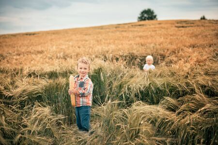 a blond boy in an orange plaid shirt and green trousers stands in a rye field, smiles and holds a wheat baguette in his hands, the baguette is partially bitten off, in the background is a second blond boy in a white T-shirtの写真素材