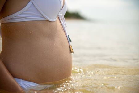 pregnant woman in a white bikini separate swimsuit is standing in the sea, swimming, pregnant belly in sea water close-upの写真素材