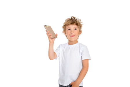 A little boy holds a phone in his hand and looks up happily with a smile. Boy stands on a white background, dressed in a white t-shirtの写真素材