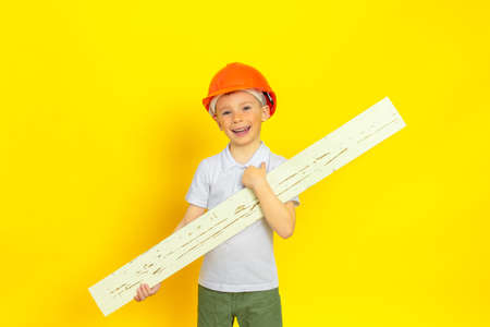 A little boy in an orange construction helmet on a yellow background, holding a wooden board in his hands, he is happy and smiling. Conception of home renovation, construction, child development, future profession.の写真素材