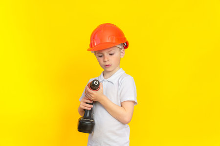 Building craft training. A boy in a kaskuke stands on a yellow background and looks attentively at a construction electric tool. The profession is a builder, the development of childrenの写真素材