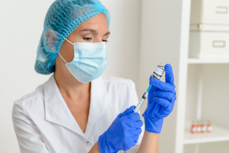 A middle-aged woman doctor picks up a syringe from an ampoule containing the covid-19 coronavirus vaccine. Close-up. Dressed in a white coat, medical gloves and a maskの写真素材