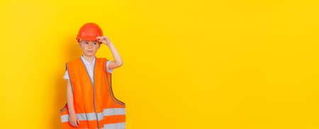 Portrait of a young (child) handsome worker or engineer wearing a helmet and orange reflective vest on a yellow background. The boy holds the helmet with his handの写真素材