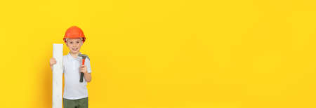Smiling child with a construction tool (hammer) and building materials stands on a yellow background, he is happy. Free space for an inscription, a bannerの写真素材