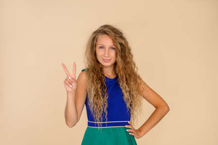 Portrait of a young beautiful girl on a beige background, smiling, looking at the camera, showing thumbs up, making a victory sign. Number twoの写真素材