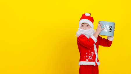 A cute Caucasian child in Santa Claus clothes and with an artificial white beard holds a round gift box high in his hands with the words New Year 2022, yellow studio backgroundの写真素材