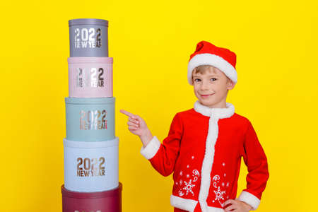 Happy smiling caucasian child stands near a tower of round gift boxes with the inscriptions New Year 2022, shows a finger at one of the boxes, yellow backgroundの写真素材