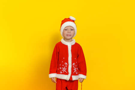 Handsome little boy stands on a yellow background dressed as Santa Claus and smiles cuteの写真素材