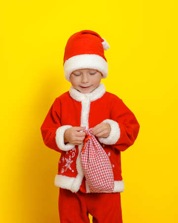 Vertical portrait of a boy dressed as Santa Claus, who is about to open a bag with gifts. Yellow backgroundの写真素材
