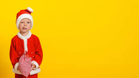 Three-year-old Caucasian boy in a Christmas Santa Claus costume holding a red in a white cage bag with gifts and squinting slyly smiles, yellow isolated backgroundの写真素材