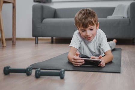 Boy-child exercising with dumbbells at home, choosing lesson on smartphone or watching having online fitness classの写真素材