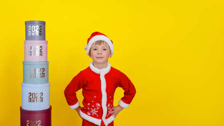A six-year-old cute child stands near a tower of round gift boxes with inscriptions New Year 2022, holds his hands at his hips, yellow background, copy spaceの写真素材