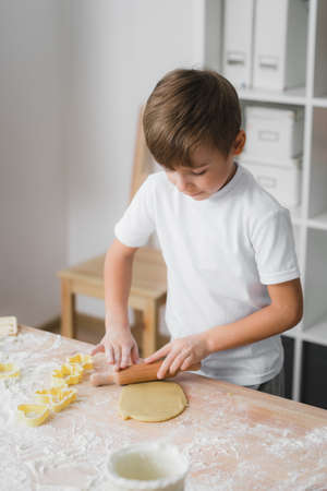 Boy rolls out raw dough with a rolling pin for making homemade baked goodsの写真素材