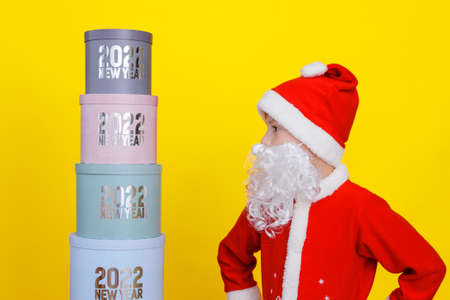 Little boy stands and looks at the tower of round gift boxes with the inscription New Year 2022, he is dressed in Santa Claus costume, yellow isolated backgroundの写真素材