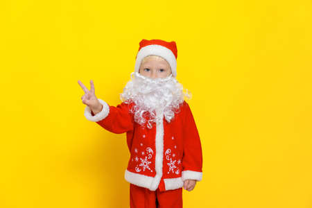 Little boy in red christmas costume with white beard shows victory gesture with hands, stands on yellow isolated backgroundの写真素材