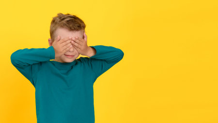 Six year old child covering eyes with hands on yellow isolated background in studioの写真素材