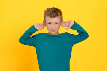 A cute boy pokes his ears with his hands in the hope of hearing better, smiles, he is in a good mood. Yellow studio backgroundの写真素材