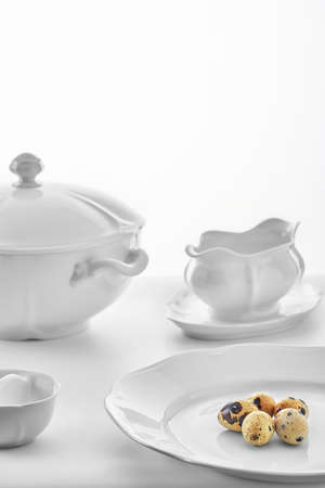 white porcelain dishware on a white background: soup tureen, gravy bowl, salt shaker and quail eggs on the large plate, high key, shallow depth of fieldの写真素材
