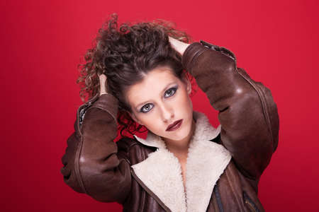 young and beautiful woman, with curly hair, holding her hair with a winter coat, on red background, studio shotの写真素材