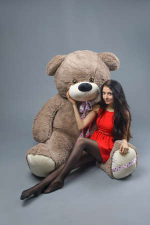 Young beautiful girl in red dress with big teddy bear soft toy happy smiling and playing on grey backgroundの写真素材