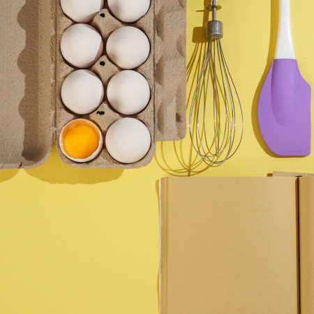 Cardboard box with white chicken eggs on a yellow background, craft paper book and pastry tools. View from above.の写真素材