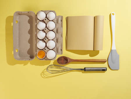 Cardboard box with white chicken eggs on a yellow background, craft paper book and pastry tools. View from above.の写真素材