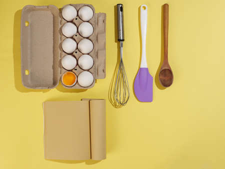 Cardboard box with white chicken eggs on a yellow background, craft paper book and pastry tools. View from above.の写真素材
