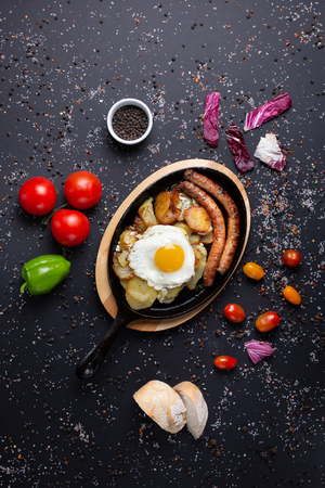 baked sausages with potatoes and fried eggs served at the restaurant,  on black background with red tomatoes, bread, red green peppers and broccoli, top viewの写真素材