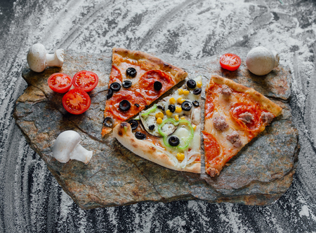 slices of different pizza, pepperoni, decorated with sliced tomatoes and mushrooms, sliced peppers,  black background on  stone with flour, Top view. Banner, close-up, copy-spaceの写真素材