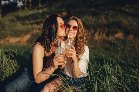 lifestyle people concept: portrait of happy girls have fun, sit down on the grass, kiss on the cheek, drink cocktail in sunglasses, at sunset, positive facial expression, outdoorの写真素材