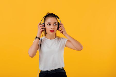 the young girl listens to the music in the headphones, dressed in a white shirt,  positive facial emotion isolated on a yellow backgroundの写真素材