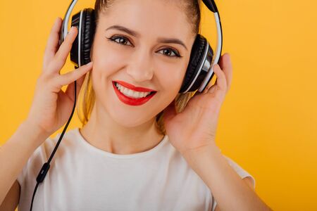 Close up. smiling girl looks at the camera with headphones on head dressed in a white shirt, positive emotional state  isolated on a yellow backgroundの写真素材