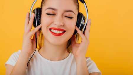 cropped. girl smiling with eyes closed, listening to music headphones on head dressed in a white shirt, positive emotional state  isolated on a yellow backgroundの写真素材