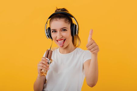 happy young girl, stick out your tongue out, headphones on head looks at camera is chocolate in hand, thumbs up dressed in a white shirt, positive emotional state  isolated on a yellow background, copy spaceの写真素材