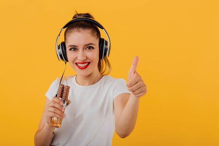 beautiful teenager, headphones on head looking at camera, chocolate in hand, keep your thumb up dressed in a white shirt, positive emotional state  isolated on a yellow background, copy spaceの写真素材