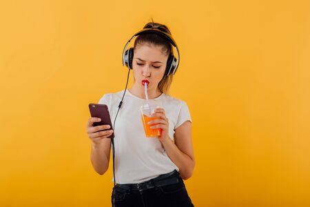 beautiful teenager, listening to music with headphones on head, looking in the phone, drink juice plastic cup, dressed in a white shirt, positive emotional state  isolated on a yellow background, copy spaceの写真素材
