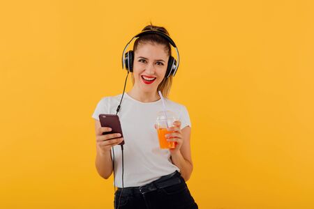 beautiful teenager, listening to music with headphones on her head, looks at the camera takes the  plastic cup with juice in hand, dressed in a white shirt, positive emotional state  isolated on a yellow background, copy spaceの写真素材