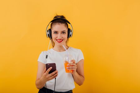 girl smiles at the camera, in her hand the phone has headphones on the head, with the juice plastic cup dressed in a white shirt,  positive facial emotion isolated on a yellow background, copy spaceの写真素材
