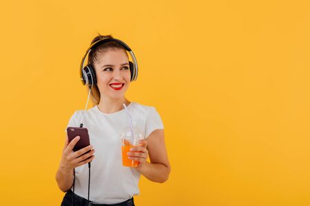 beautiful girl smiles, in her hand the phone has headphones on the head with the juice plastic cup dressed in a white shirt,  positive facial emotion isolated on a yellow background, copy spaceの写真素材