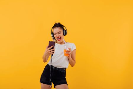 the beautiful girl smiles, the smartphone has headphones on the head, with the juice  plastic cup, makes the selfie dressed in a white shirt,  positive facial emotion isolated on a yellow background, copy spaceの写真素材