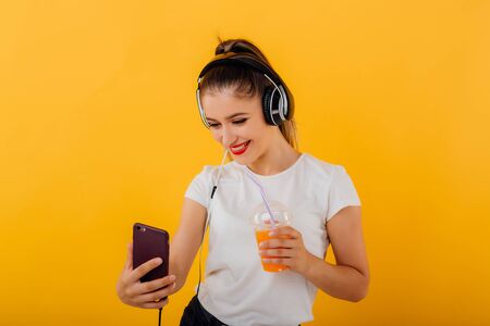 beautiful girl smiles and makes the selfie, in the smartphone hand has headphones with the juice  plastic cup, dressed in a white shirt,  positive facial emotion isolated on a yellow background, copy spaceの写真素材